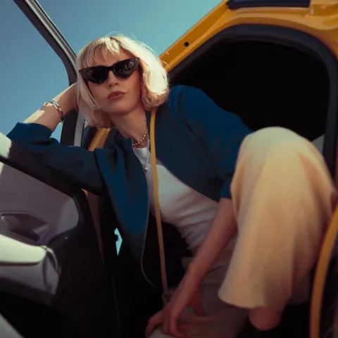 A lady hanging out of a Ford Capri with black sunglasses and a bag hanging from her neck.